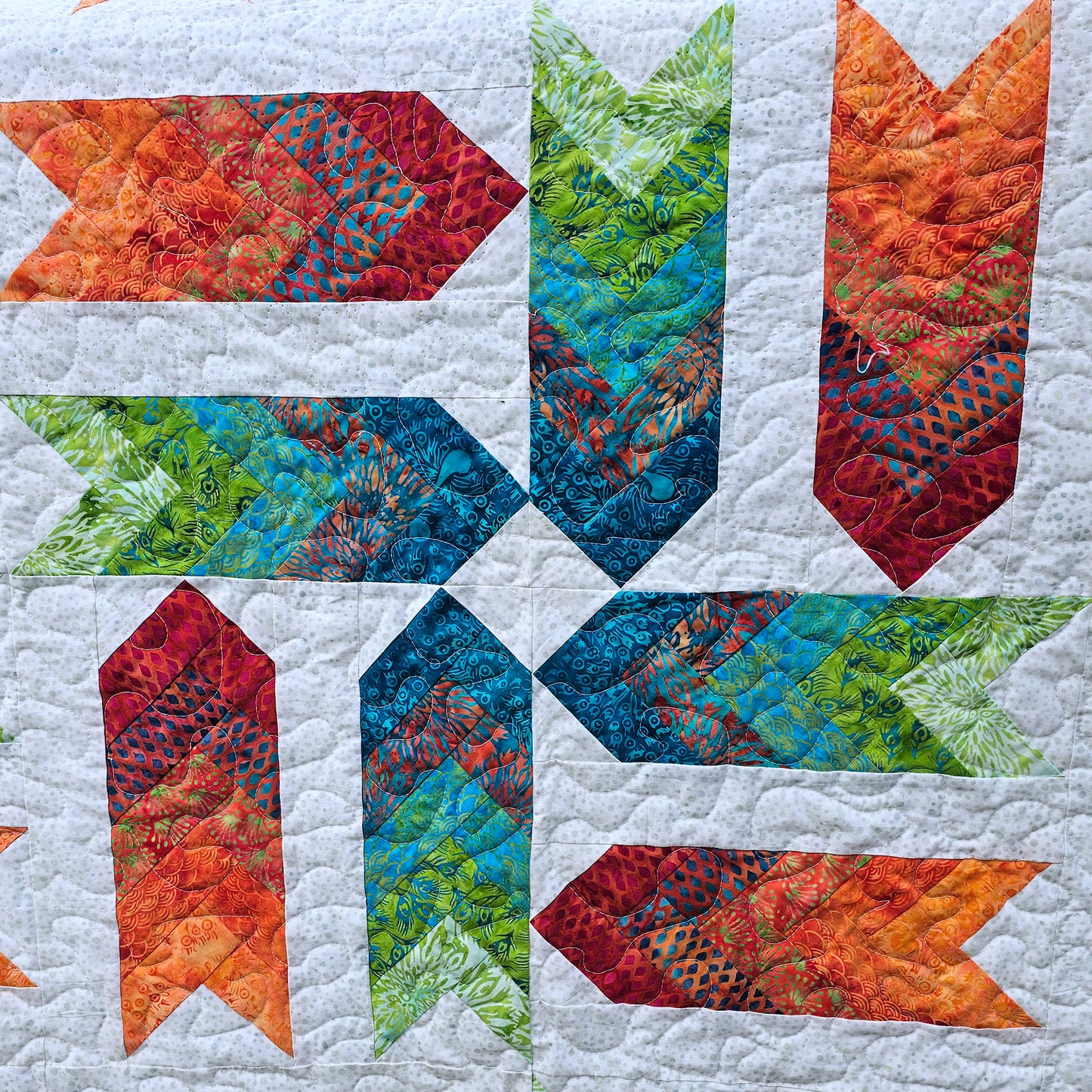 A close-up of a quilted fabric block pattern featuring six arrow-shaped patches arranged in a pinwheel. The patches display a multicolor gradient of red, orange, yellow, green, and blue on a white quilted background.