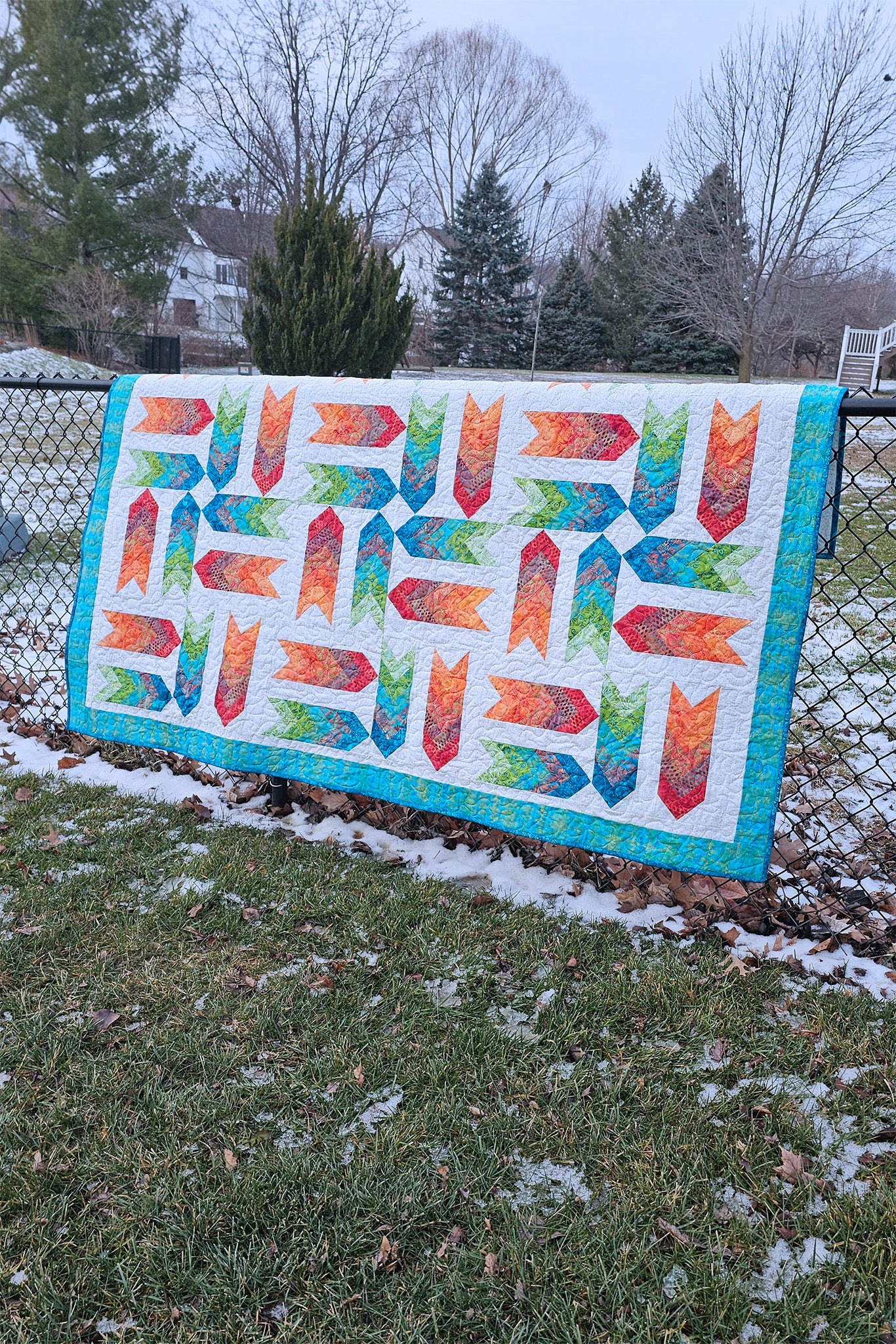 Fletching, a colorful quilted blanket or small quilt hanging outdoors on a chain-link fence in a grassy yard with patches of snow and trees in the background.