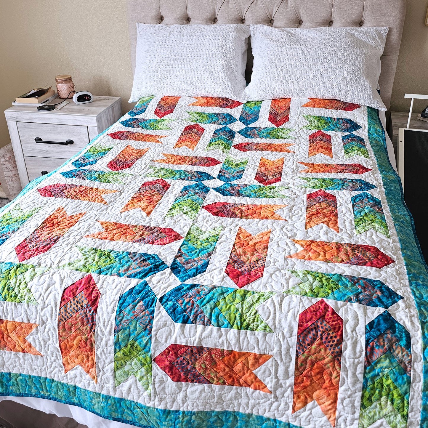 A colorful quilted bedspread with a geometric patchwork pattern styled on a bed with white pillows and a tufted headboard.