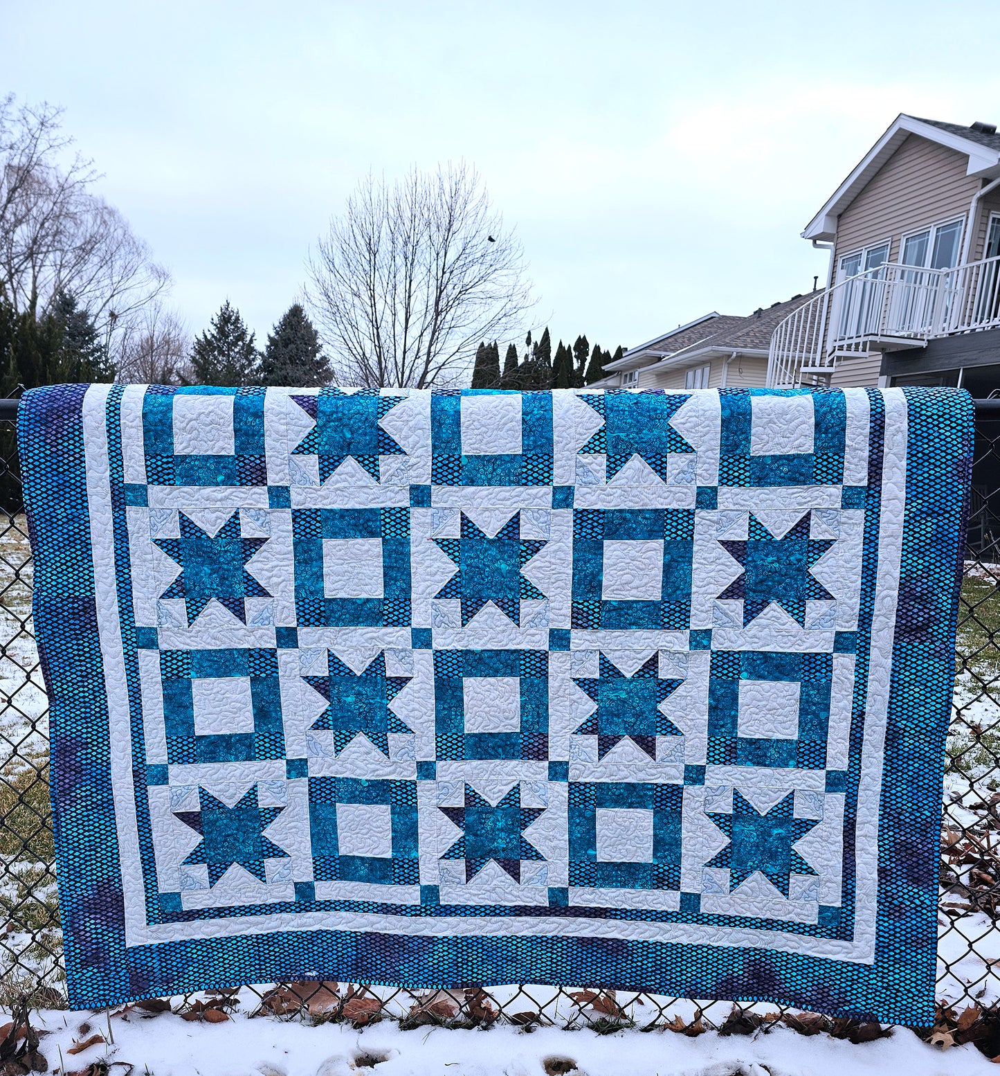 A square quilted blanket with a geometric star and square pattern in teal blue and white, draped over a chain-link fence in an outdoor backyard setting with snow on the ground and houses in the background.