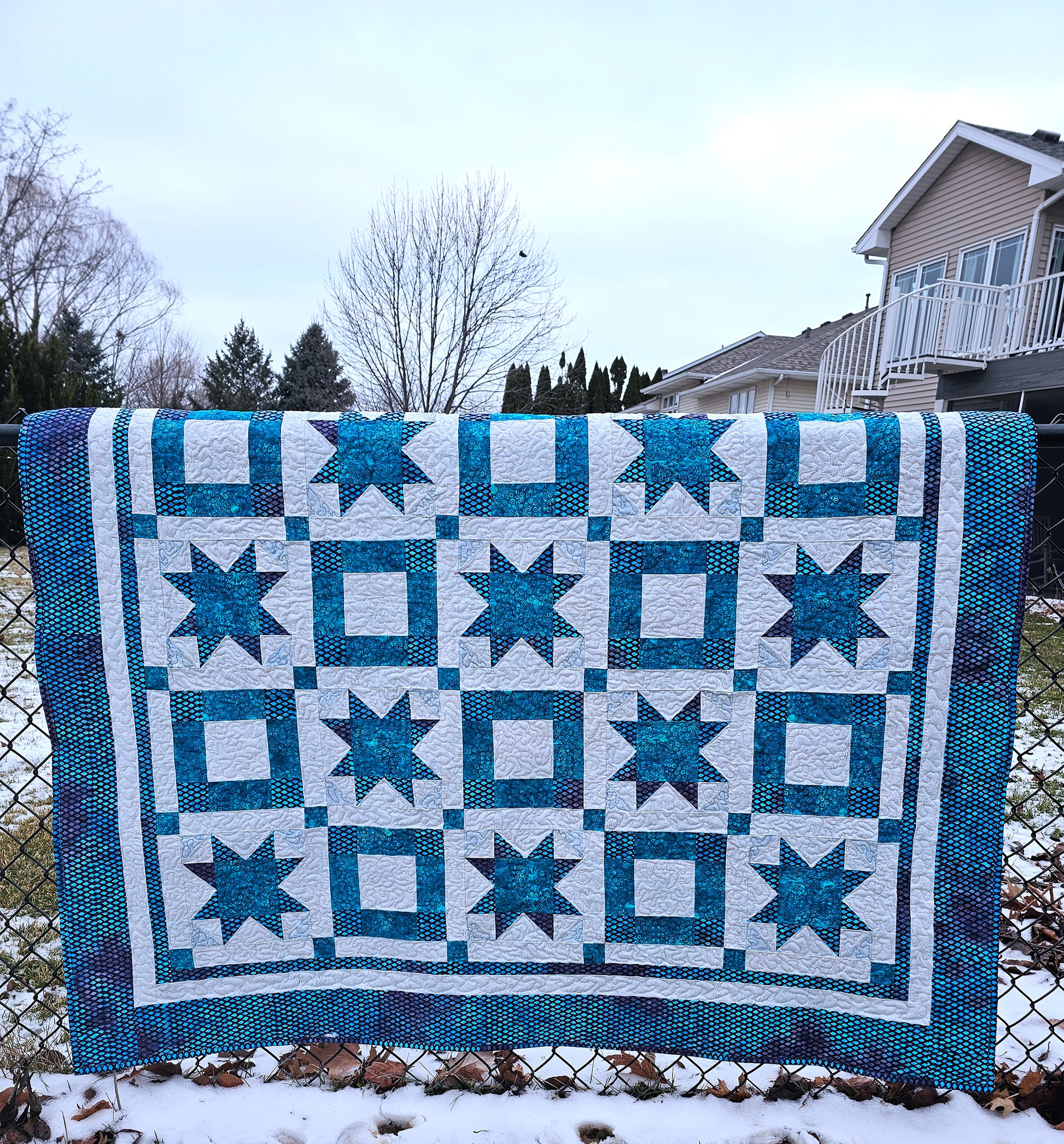 A square quilted blanket with a geometric star and square pattern in teal blue and white, draped over a chain-link fence in an outdoor backyard setting with snow on the ground and houses in the background.